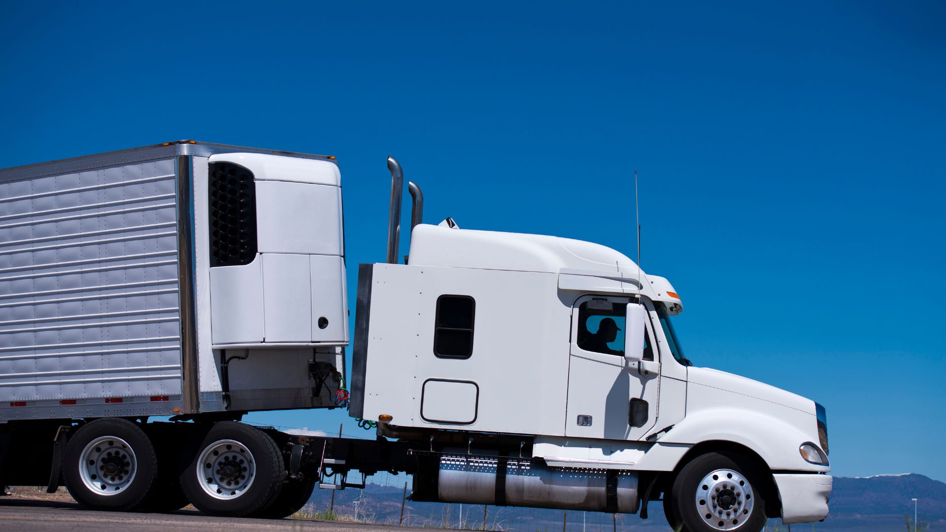 White semi-truck with trailer against bright blue sky and distant mountains