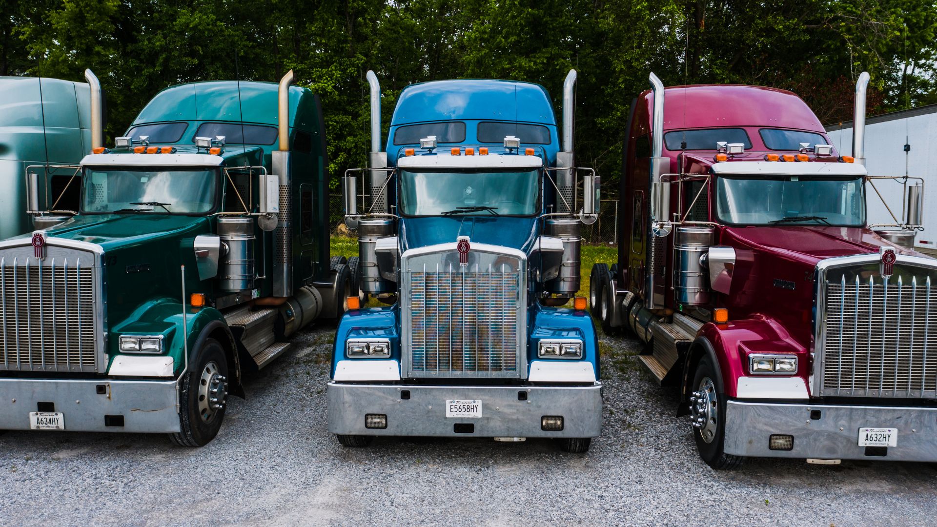 Three semi-trucks in teal, blue, and red parked on gravel lot
